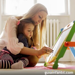 una mujer juega con una niña pequeña a escribir en una pizarra, mientras la tiene en sus piernas