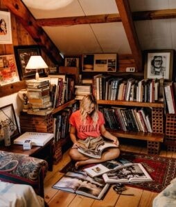 una joven, de unos veinte años, está sentada en un piso de madera, apoyada en un estante hecho con tablones y ladrillos, lleno de libros y cuadros, en la parte de arriba de un chalet. TIene una lámpara de noche pequeña, y está sobre una alfombra persa y un piso de madera. Ve libros de fotografías, y se ve feliz en un lugar acogedor y cálido