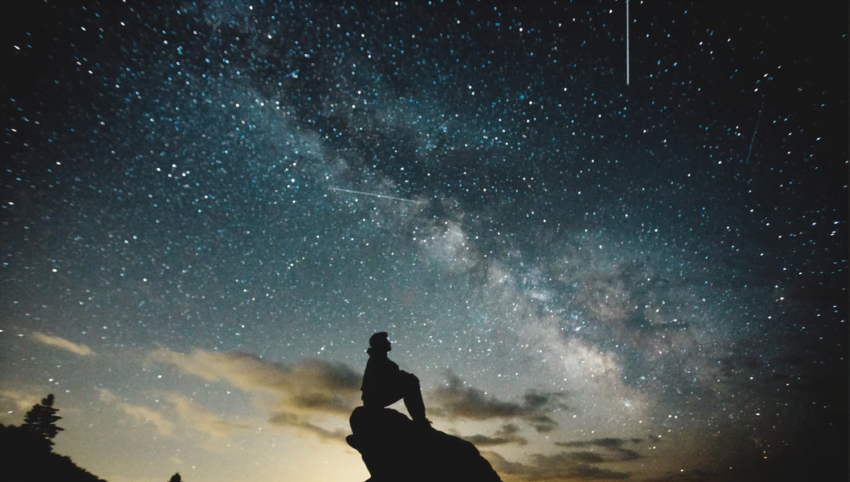 una persona diminuta, sentada en una roca, disfruta de la inmensidad del cielo nocturno, tachonado de estrellas