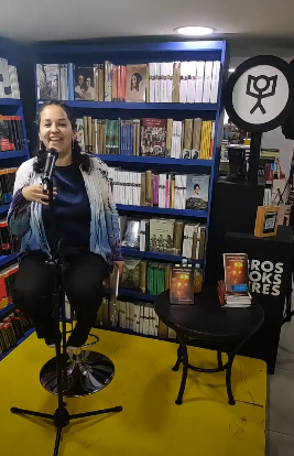yo, en un pequeño foro, entre anaqueles de libros, sentada en un banquito, con el micrófono enfrente, con cara de que me la estoy pasando de lo lindo en el espectáculo