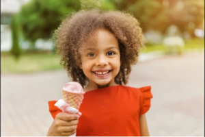 una niña afrodescendiente, con hermosos cabellos rizados y desmelenados, sostiene un helado de fresa con la sonrisa feliz que sólo una niña puede tener